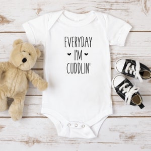 May include: White baby onesie with black text that reads "EVERYDAY I'M CUDDLIN'" with two small hearts above the word "I'M".