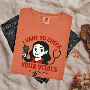 May include: A terracotta-colored t-shirt featuring a graphic of a cartoon vampire with fangs, holding a reflex hammer and wearing a stethoscope. The text reads I VANT TO CHECK YOUR VITALS in red lettering. The shirt has a Comfort Colors brand label.