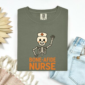 May include: An olive green t-shirt featuring a graphic of a cartoon skeleton wearing a nurse cap with a red cross. The skeleton is waving, and the text below reads BONE-AFIDE NURSE in orange. The shirt has a Comfort Colors brand neck label.