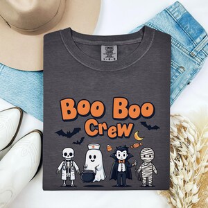 May include: A charcoal gray t-shirt featuring the orange text "Boo Boo Crew" above a graphic of a skeleton doctor, a ghost nurse, a vampire, and a mummy. The shirt has a Comfort Colors label at the inner neckline and is displayed on a flat surface.