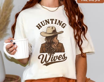 May include: Cream-colored t-shirt featuring a graphic of a woman wearing a cowboy hat and jacket, with the words "HUNTING WIVES" in a brown, vintage font. The design has a rustic, outdoorsy theme.