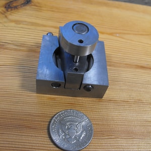 May include: A small, silver metal device with a cylindrical top and a square base. A U.S. half-dollar coin is next to the device. The device is on a wooden surface.
