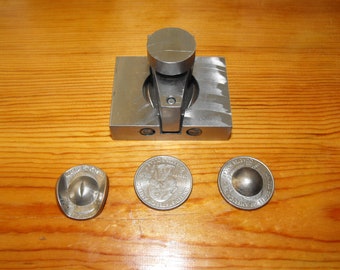 Puede incluir: Una herramienta metálica para dar forma a monedas con una base cuadrada y una parte superior cilíndrica, junto a tres monedas de plata. Una moneda es plana, las otras tienen forma de cúpula y de ojo. Los objetos están sobre una superficie de madera.
