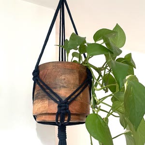 May include: A black macrame plant hanger with a wooden pot and a green plant hanging from the ceiling.