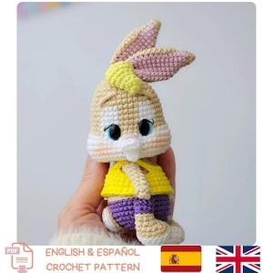 Peut inclure: Une poupée lapin au crochet avec un corps beige, des oreilles doublées de rose et un gilet jaune sur un short violet. Le lapin a des yeux noirs et un museau blanc. Le texte "ENGLISH & ESPAÑOL CROCHET PATTERN" est inclus.
