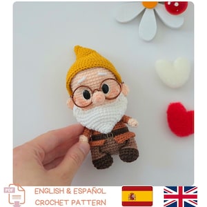 Puede incluir: Un pequeño muñeco gnomo de ganchillo con un gorro puntiagudo de color amarillo mostaza, una espesa barba blanca y gafas redondas de montura marrón. La figura lleva una túnica marrón con un pequeño cinturón y pantalones de color marrón oscuro.