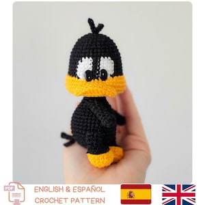 Puede incluir: Un juguete de patito tejido a crochet en negro, amarillo y blanco. El patito tiene ojos blancos grandes con pupilas negras, un pico amarillo y pies amarillos. El patrón de crochet está disponible en inglés y español.