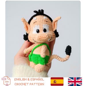 Puede incluir: Muñeco de ganchillo con piel clara, pelo negro y orejas grandes. El muñeco lleva un pantalón corto verde y tiene una cola larga. El texto "ENGLISH & ESPAÑOL CROCHET PATTERN" es visible.