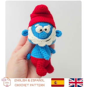 Puede incluir: Un muñeco de los Pitufos tejido a ganchillo con gorro y pantalón rojos, cuerpo azul y barba blanca. El muñeco está hecho a mano y presenta costuras detalladas. La imagen también incluye el texto "ENGLISH & ESPAÑOL CROCHET PATTERN".
