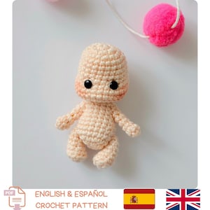 Könnte beinhalten: Eine gehäkelte Puppe in hellem Pfirsichton mit schwarzen Augen und rosigen Wangen. Die Puppe hat kleine Arme und Beine und sitzt. Das Bild zeigt auch einen rosa Pompon an einer Schnur und den Text "ENGLISH & ESPAÑOL CROCHET PATTERN".