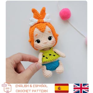 Puede incluir: Muñeca de ganchillo hecha a mano de un personaje de dibujos animados con pelo naranja y un lazo blanco. La muñeca lleva un top verde y pantalones cortos azules. La imagen incluye el texto "ENGLISH & ESPAÑOL CROCHET PATTERN".