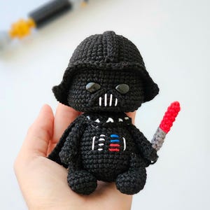 May include: A black crochet Darth Vader doll with a helmet, cape, and a red and gray lightsaber. The doll has white details on the chest and face. The doll is held in a hand.