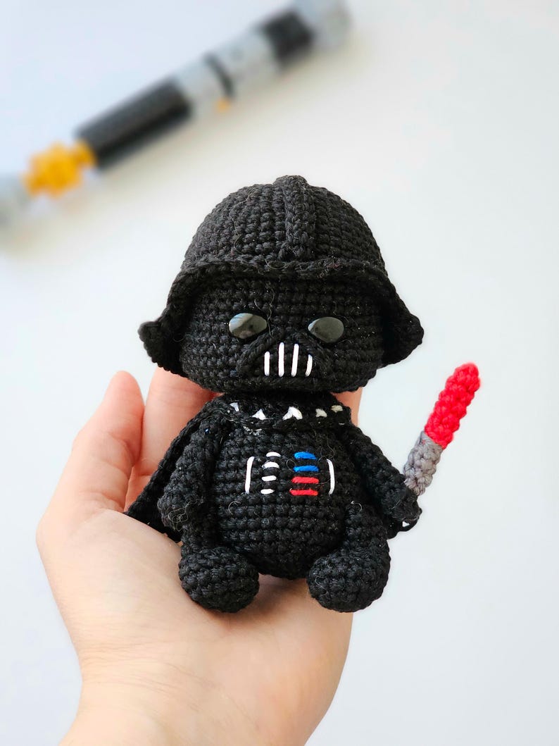 May include: A black crochet Darth Vader doll with a helmet, cape, and a red and gray lightsaber. The doll has white details on the chest and face. The doll is held in a hand.