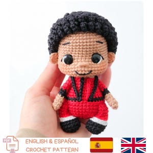 May include: A small amigurumi doll featuring a head with curly black yarn hair, tan skin, and large black plastic eyes. The doll wears a red jacket with black trim, red pants, white cuffs, and black shoes, all crafted from crocheted yarn.