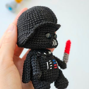 May include: A handmade black crochet doll resembling Darth Vader, complete with a helmet, mask, and a red and gray lightsaber. The doll has white, red, and blue accents on the chest. The doll is approximately 4 inches tall.