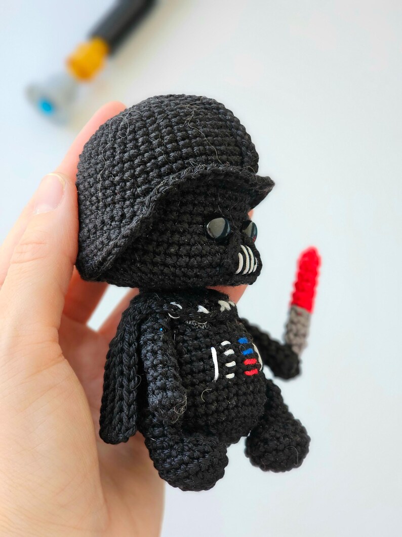 May include: A handmade black crochet doll resembling Darth Vader, complete with a helmet, mask, and a red and gray lightsaber. The doll has white, red, and blue accents on the chest. The doll is approximately 4 inches tall.