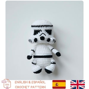 以下が含まれることがあります： 白と黒の編みぐるみのストームトルーパー人形。人形は、黒いアクセント、黒い目、胸の黒いストライプを備えた詳細なヘルメットデザインが特徴です。かぎ針編みのパターンです。