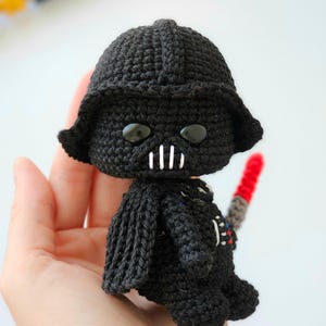 May include: A handmade black crochet Darth Vader doll with a red lightsaber. The doll features a detailed helmet, cape, and iconic facial features. The doll is approximately 4 inches tall.
