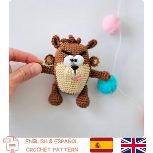 Może przedstawiać: Szydełkowa figurka Diabła Tasmańskiego w kolorach brązu, beżu i różu, z niebieskim pomponem. Figurka ma psotny wyraz twarzy z wystającym językiem. Obraz zawiera tekst "ENGLISH & ESPAÑOL CROCHET PATTERN".