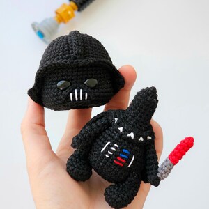 May include: Handmade crochet Darth Vader doll and helmet in black yarn. The doll has a red and gray lightsaber. The helmet has black eyes and a white mouth. A gray and yellow toy lightsaber is in the background.