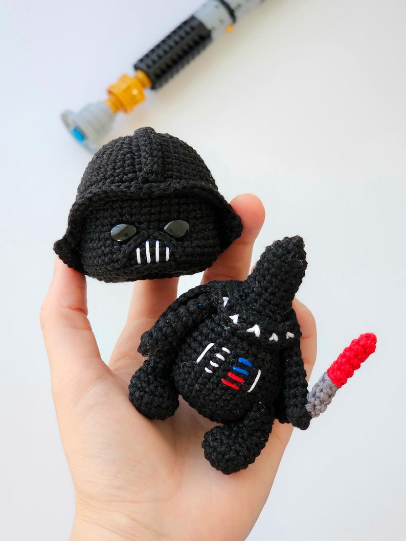 May include: Handmade crochet Darth Vader doll and helmet in black yarn. The doll has a red and gray lightsaber. The helmet has black eyes and a white mouth. A gray and yellow toy lightsaber is in the background.