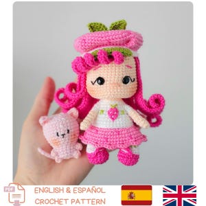 Puede incluir: Una muñeca de ganchillo hecha a mano con pelo rosa, boina a juego y un vestido blanco con diseño de fresa. Un pequeño ratón rosa de ganchillo está al lado de la muñeca. El texto "ENGLISH & ESPAÑOL CROCHET PATTERN" está en la parte inferior.