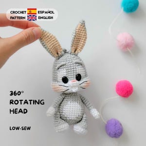 Könnte beinhalten: Eine gehäkelte Hasenpuppe in Grau und Weiß mit langen Ohren, schwarzen Augen und rosa Wangen. Der Text lautet: CROCHET PATTERN ESPAÑOL ENGLISH, 360° ROTATING HEAD und LOW-SEW. Der Hase hat einen hellgrauen Körper mit einem weißen Bauchfleck.
