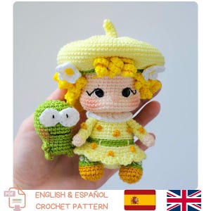 Puede incluir: Una muñeca de ganchillo hecha a mano y una pequeña rana verde. La muñeca lleva un vestido amarillo, un sombrero y tiene el pelo rizado amarillo. La rana tiene grandes ojos blancos. El texto "ENGLISH & ESPAÑOL CROCHET PATTERN" está en la parte inferior.