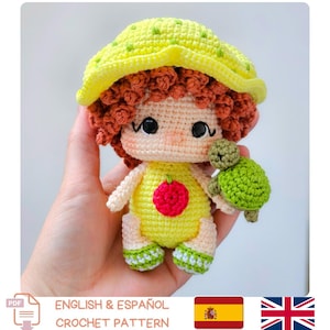 Puede incluir: Una muñeca de ganchillo hecha a mano con cabello rizado rojo, que lleva un atuendo amarillo con una manzana roja y un sombrero amarillo. La muñeca sostiene una tortuga verde. La imagen incluye el texto "ENGLISH & ESPAÑOL CROCHET PATTERN".