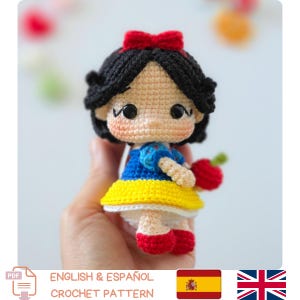 Puede incluir: Una pequeña muñeca de ganchillo con cabello negro y un lazo rojo, un corpiño azul con mangas abullonadas y una falda amarilla. La muñeca tiene grandes ojos negros, mejillas sonrosadas y sostiene una manzana roja. Texto: English & Español Crochet Pattern.