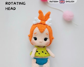 May include: A crochet doll with bright orange hair styled with a white bone accessory, wearing a lime green top with black spots and blue bottoms. Text reads 360 ROTATING HEAD, CROCHET PATTERN, ESPAÑOL, and ENGLISH. Pink pom-poms are visible.