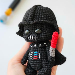 May include: A handmade black crochet Darth Vader doll with a red and gray lightsaber. The doll has a black helmet, a white mouth, and a chest plate with red, blue, and white details. The doll is approximately 4 inches tall.