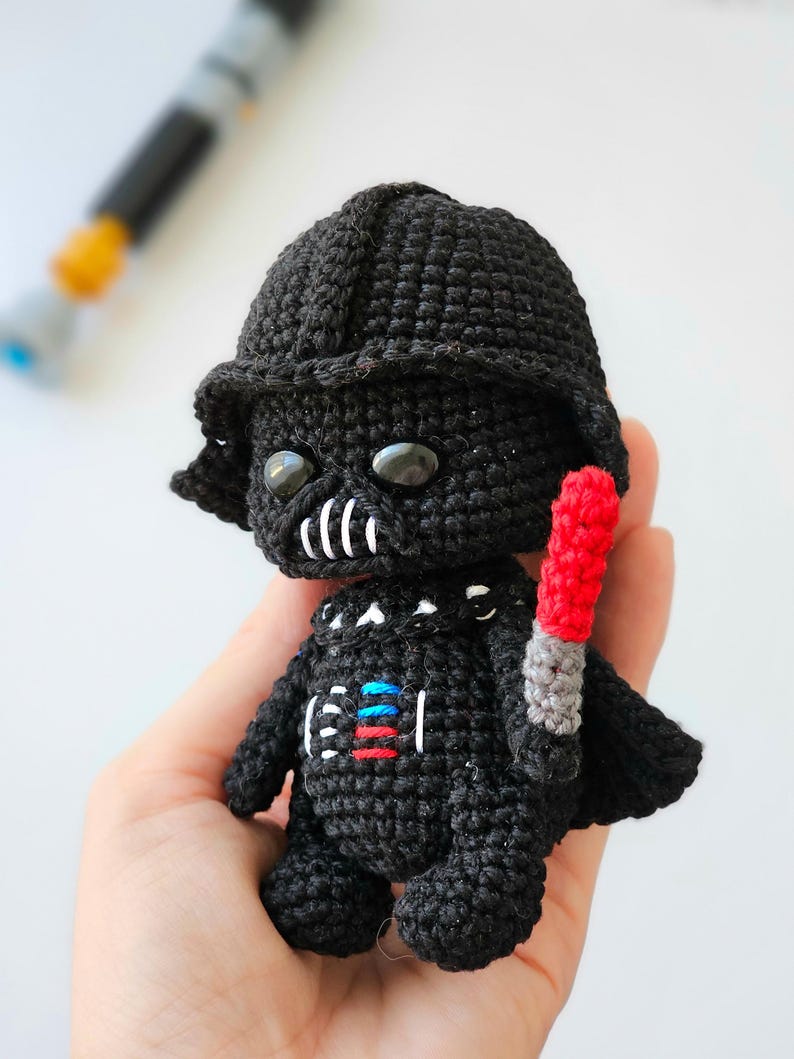 May include: A handmade black crochet Darth Vader doll with a red and gray lightsaber. The doll has a black helmet, a white mouth, and a chest plate with red, blue, and white details. The doll is approximately 4 inches tall.
