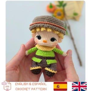 May include: A small crocheted doll featuring a tan face with black eyes, a light green shirt, dark brown pants, and a brown hat with an orange trim. The doll has yellow hair and a yellow collar, with a small orange nose and pink blush on the cheeks.
