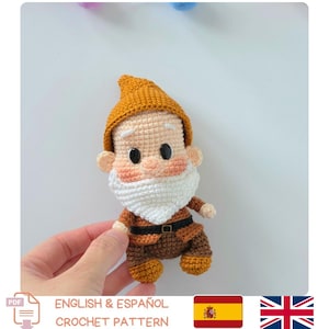 May include: A small, handcrafted crochet gnome doll featuring a mustard yellow pointed hat, a white beard, and a brown tunic with a black belt. The doll has a light peach-colored face with black plastic eyes, rosy cheeks, and white eyebrows.