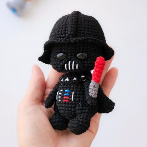May include: Crocheted Darth Vader doll in black with a red and gray lightsaber. The doll has a black helmet, a detailed chest plate, and black eyes. A Star Wars themed handmade item.