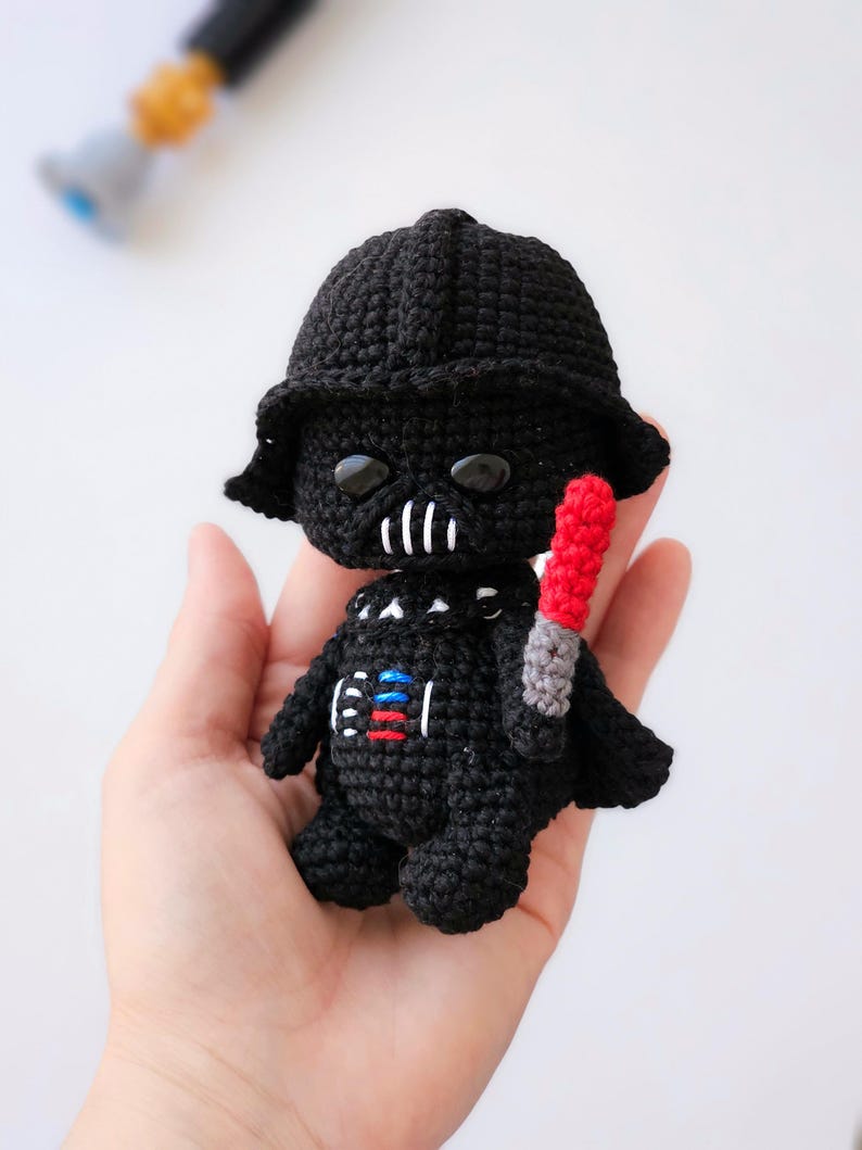 May include: Crocheted Darth Vader doll in black with a red and gray lightsaber. The doll has a black helmet, a detailed chest plate, and black eyes. A Star Wars themed handmade item.