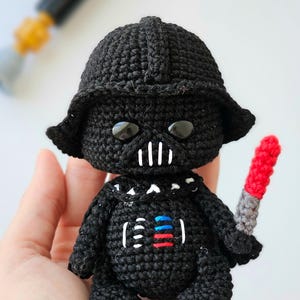 May include: A handmade black crochet doll resembling Darth Vader, complete with a helmet, mask, and a red and gray lightsaber. The doll has white accents on the mask and chest, and large black eyes. The overall design is a cute, stylized representation.