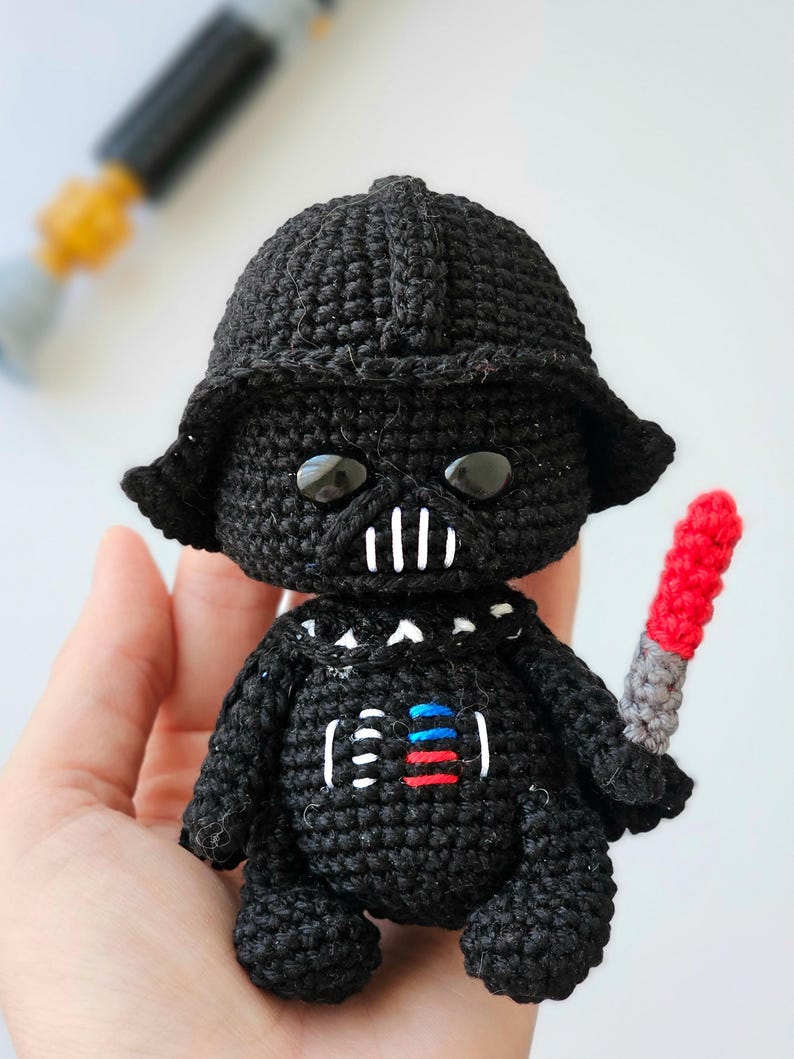 May include: A handmade black crochet doll resembling Darth Vader, complete with a helmet, mask, and a red and gray lightsaber. The doll has white accents on the mask and chest, and large black eyes. The overall design is a cute, stylized representation.