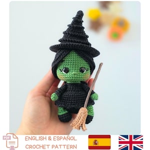 May include: A small green-skinned witch doll made of crocheted yarn. The doll wears a black pointed hat and a black dress, featuring large black eyes and pink blush on the cheeks. It holds a small tan-colored broomstick made of yarn.