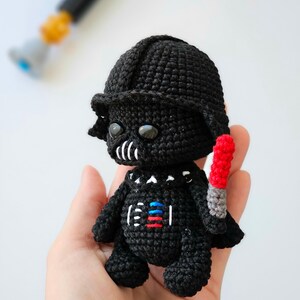 May include: A handmade black crochet figure of Darth Vader, complete with helmet and a red and gray lightsaber. The figure has white and colored details on the chest. The figure is held in a hand, with a blurred lightsaber in the background.