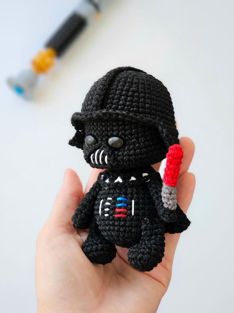 May include: A handmade black crochet figure of Darth Vader, complete with helmet and a red and gray lightsaber. The figure has white and colored details on the chest. The figure is held in a hand, with a blurred lightsaber in the background.