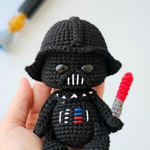 May include: A black crocheted Darth Vader doll with a red and gray lightsaber. The doll has a black helmet, a white mouth, and a chest design with red, white, and blue accents. The doll is handmade.