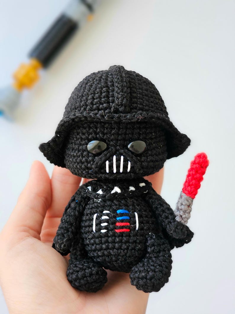 May include: A black crocheted Darth Vader doll with a red and gray lightsaber. The doll has a black helmet, a white mouth, and a chest design with red, white, and blue accents. The doll is handmade.
