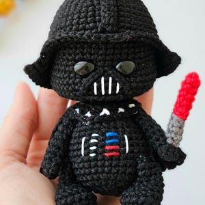 May include: A handmade, crocheted Darth Vader doll in black yarn. The doll features a helmet, mask, and details like the chest panel with red, blue, and white accents. It holds a red and gray lightsaber. The doll is approximately 4 inches tall.