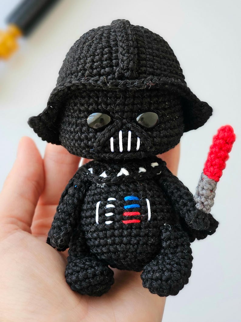 May include: A handmade, crocheted Darth Vader doll in black yarn. The doll features a helmet, mask, and details like the chest panel with red, blue, and white accents. It holds a red and gray lightsaber. The doll is approximately 4 inches tall.