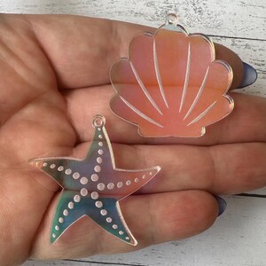 May include: Two iridescent acrylic charms: a starfish and a seashell. The starfish is multi-colored with white dots. The seashell is orange and pink with white lines. Both charms have a small hole for hanging.
