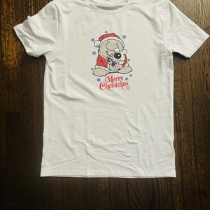 May include: White t-shirt with a cartoon image of a bear wearing a Santa hat and holding a smaller bear. The text "Merry Christmas" is printed below the image.