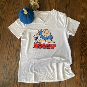 May include: White t-shirt with a cartoon illustration of a man and a dog. The man is wearing a blue shirt and the text "Ziggy" is printed in red below the illustration.