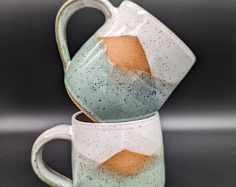May include: Two handmade ceramic mugs stacked on top of each other. The mugs have a rounded shape with a handle. The mugs are white, speckled with brown and green, and feature a geometric design in shades of brown and green.